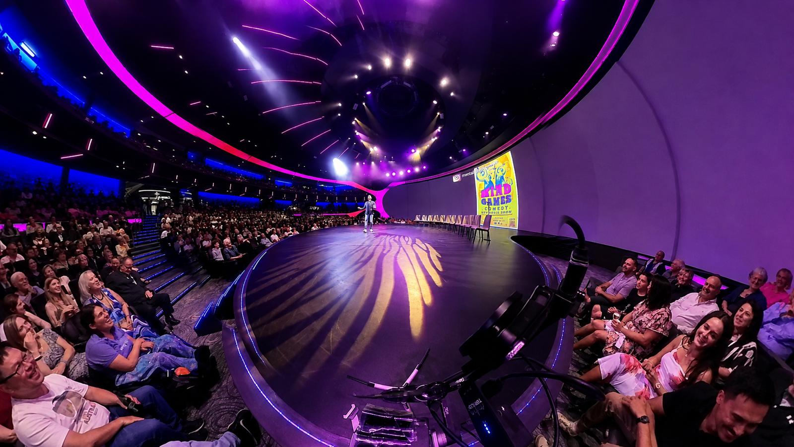 Wide view of a packed cruise-ship theatre with Phoenix alone on stage beside the Mind Games comedy hypnosis show screen