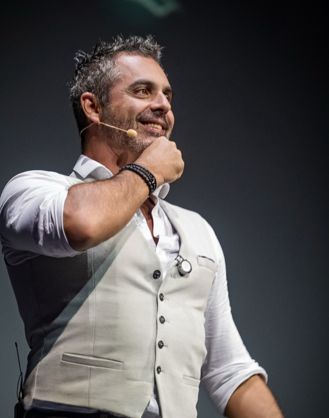 Phoenix on stage in a waistcoat and headset microphone, hand to his chin, smiling at the audience
